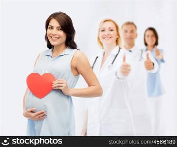 pregnancy, healthcare, medicine, people and expectation concept - happy pregnant woman with red heart shape touching her belly over group of doctors or obstetricians showing thumbs up background