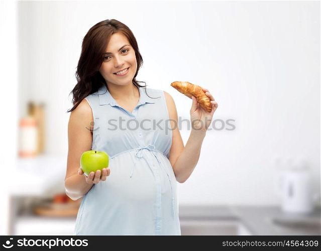 pregnancy, healthy eating, junk food and people concept - happy pregnant woman choosing between green apple and croissant over home kitchen room background