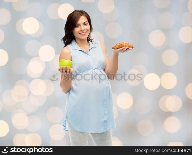 pregnancy, healthy eating, junk food and people concept - happy pregnant woman choosing between green apple and croissant over holidays lights background