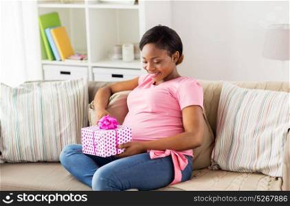 pregnancy, holidays and people concept - happy pregnant african american woman sitting on sofa with gift box at home. happy african american pregnant woman with gift