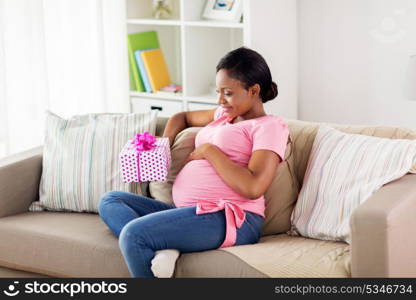 pregnancy, holidays and people concept - happy pregnant african american woman sitting on sofa with gift box at home. happy african american pregnant woman with gift