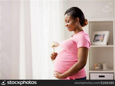 pregnancy, holidays and people concept - happy pregnant african american woman with flower looking through window at home. happy african american pregnant woman with flower