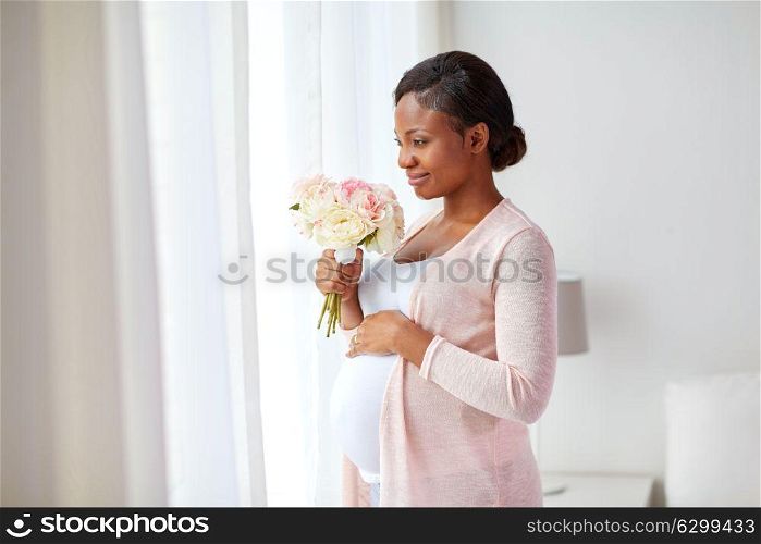 pregnancy, holidays and people concept - happy pregnant african american woman with flowers looking through window at home. happy african american pregnant woman with flowers