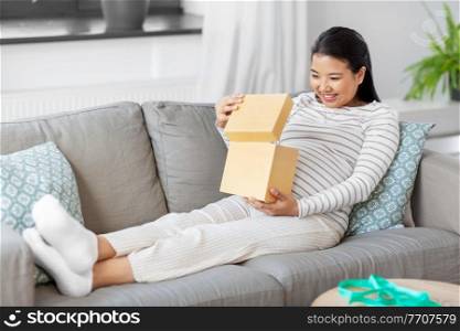 pregnancy, holidays and people concept - happy smiling pregnant asian woman opening gift box sitting on sofa at home. happy pregnant woman opening gift box at home