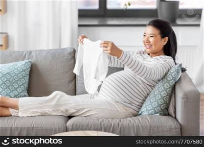 pregnancy, holidays and people concept - happy smiling pregnant asian woman with baby’s bodysuit at home. happy pregnant woman with baby’s bodysuit at home