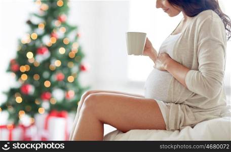 pregnancy, holidays, drinks, people and expectation concept - close up of happy pregnant woman with cup drinking tea sitting on bed at home bedroom over christmas tree background