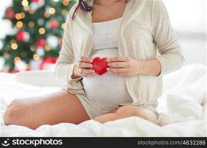 pregnancy, holidays, love, people and expectation concept - close up of pregnant woman with red heart in bed over christmas tree background