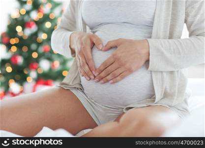 pregnancy, holidays, love, people and expectation concept - close up of pregnant woman making heart gesture in bed at home over christmas tree background