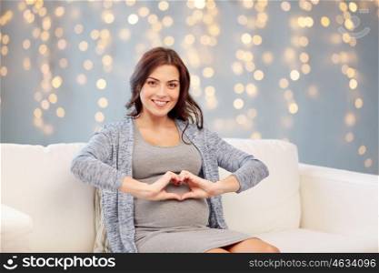 pregnancy, holidays, love, people and expectation concept - happy pregnant woman sitting on sofa and making heart gesture over christmas lights background