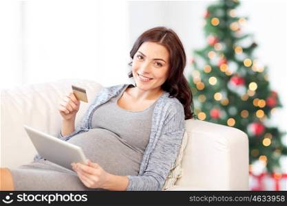 pregnancy, holidays, online shopping, technology and people concept - happy pregnant woman with tablet pc computer and credit card on couch over christmas tree background