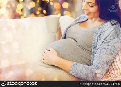 pregnancy, holidays, people and expectation concept - close up of happy pregnant woman with big belly holding cup and drinking tea over christmas lights background