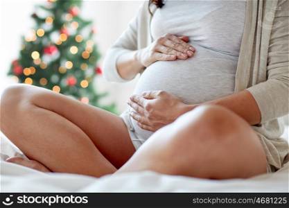pregnancy, holidays, people and expectation concept - close up of pregnant woman sitting in bed and touching her belly at home over christmas tree background