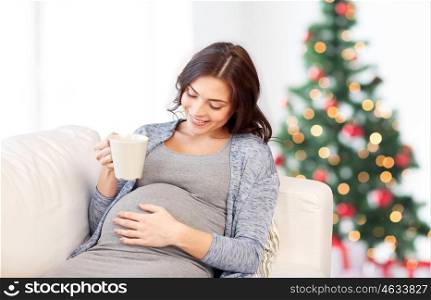 pregnancy, holidays, people and expectation concept - happy pregnant woman with cup drinking tea on sofa over christmas tree background