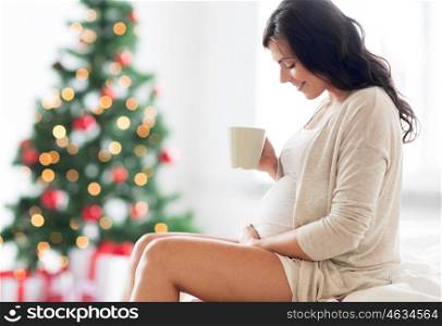 pregnancy, holidays, people and expectation concept - happy pregnant woman with cup drinking tea sitting on bed over christmas tree background