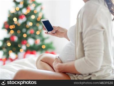 pregnancy, holidays, technology, people and expectation concept - close up of pregnant woman with smartphone in bed over christmas tree background