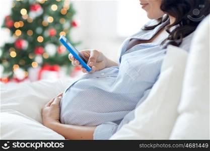pregnancy, holidays, technology, people and expectation concept - close up of pregnant woman with smartphone in bed at home over christmas tree background