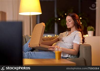 pregnancy, leisure and people concept - happy smiling pregnant woman on sofa watching tv and eating takeaway pizza at home. pregnant woman with pizza box watching tv at home
