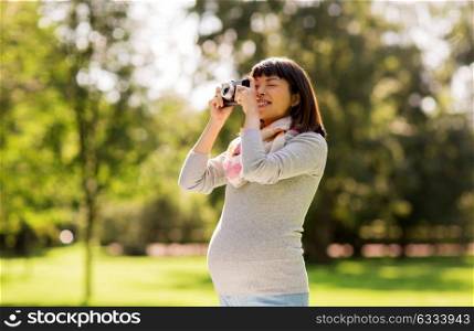 pregnancy, leisure and photography concept - happy pregnant asian woman with film camera photographing at park. happy pregnant asian woman with camera at park
