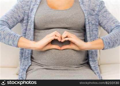 pregnancy, love, care, people and expectation concept - close up of happy pregnant woman making heart gesture at home