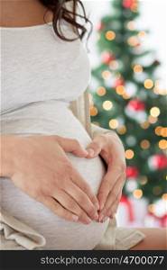 pregnancy, love, holidays, people and expectation concept - close up of pregnant woman making heart gesture at home over christmas tree background
