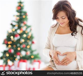 pregnancy, love, holidays, people and expectation concept - happy pregnant woman making heart gesture at home over christmas tree background