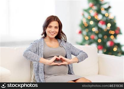 pregnancy, love, holidays, people and expectation concept - happy pregnant woman sitting on sofa and making heart gesture over christmas tree background
