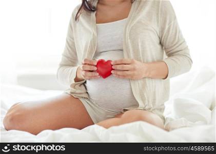 pregnancy, love, people and expectation concept - close up of pregnant woman with red heart in bed at home bedroom
