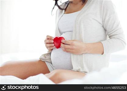 pregnancy, love, people and expectation concept - close up of pregnant woman with red heart in bed at home bedroom