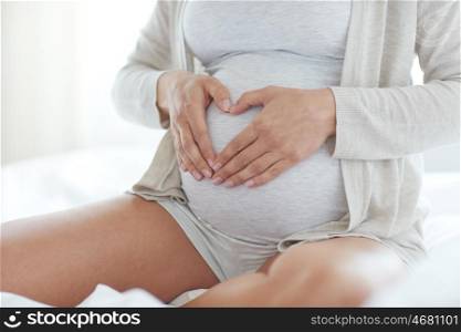 pregnancy, love, people and expectation concept - close up of pregnant woman making heart gesture in bed at home