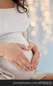 pregnancy, love, people and expectation concept - close up of pregnant woman making heart gesture over holidays lights background
