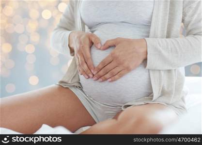 pregnancy, love, people and expectation concept - close up of pregnant woman making heart gesture in bed over holidays lights background