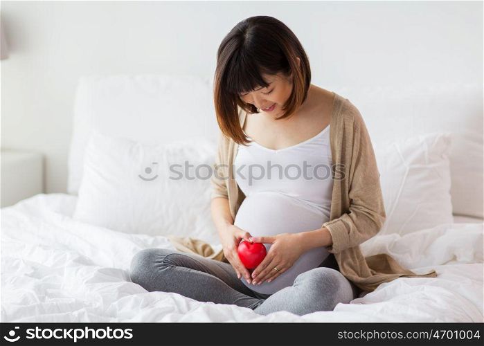 pregnancy, love, people and expectation concept - happy pregnant asian woman sitting in bed at home and holding small red heart. happy pregnant asian woman with red heart in bed. happy pregnant asian woman with red heart in bed