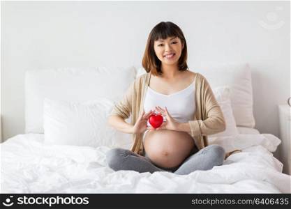 pregnancy, love, people and expectation concept - happy pregnant asian woman sitting in bed with red heart at home. happy pregnant asian woman with red heart in bed