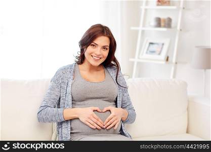 pregnancy, love, people and expectation concept - happy pregnant woman sitting on sofa and making heart gesture at home