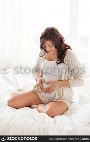 pregnancy, love, people and expectation concept - happy pregnant woman sitting on sofa and making heart gesture at home bedroom