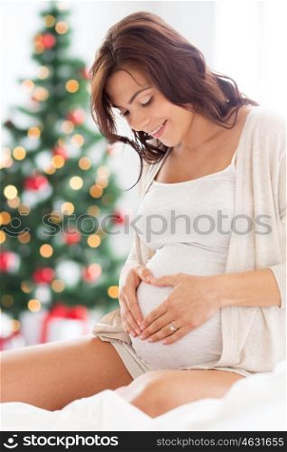 pregnancy, love, people and expectation concept - happy pregnant woman sitting on bed and making heart gesture over christmas tree background