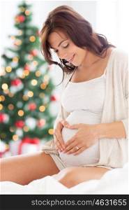 pregnancy, love, people and expectation concept - happy pregnant woman sitting on bed and making heart gesture over christmas tree background