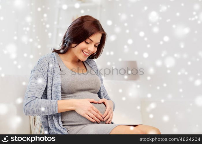 pregnancy, love, people and expectation concept - happy pregnant woman sitting on sofa and making heart gesture at home over snow