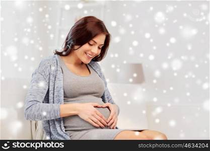 pregnancy, love, people and expectation concept - happy pregnant woman sitting on sofa and making heart gesture at home over snow