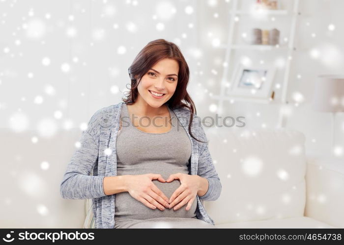 pregnancy, love, people and expectation concept - happy pregnant woman sitting on sofa and making heart gesture at home over snow