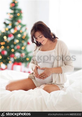 pregnancy, love, people and expectation concept - happy pregnant woman sitting on sofa and making heart gesture over christmas tree background
