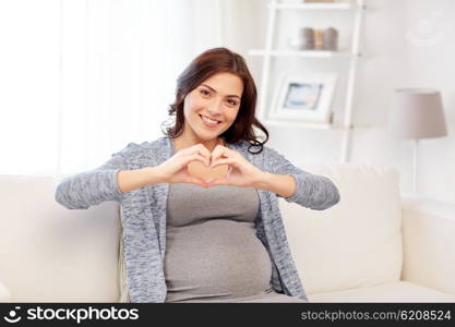 pregnancy, love, people and expectation concept - happy pregnant woman sitting on sofa and making heart gesture at home