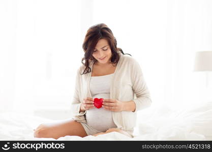 pregnancy, love, people and expectation concept - happy pregnant woman with red heart in bed at home bedroom