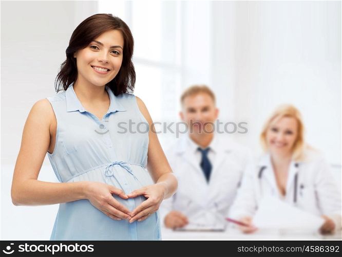 pregnancy, love, people, medicine and fertility concept - happy pregnant woman making heart gesture on belly over medics at maternity hospital background