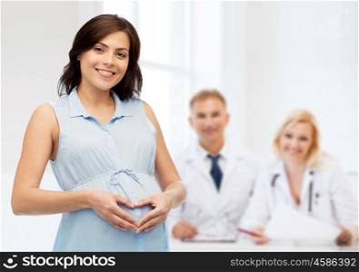 pregnancy, love, people, medicine and fertility concept - happy pregnant woman making heart gesture on belly over medics at maternity hospital background