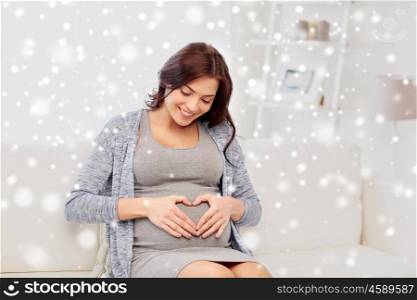 pregnancy, love, people, winter and expectation concept - happy pregnant woman sitting on sofa and making heart gesture at home over snow