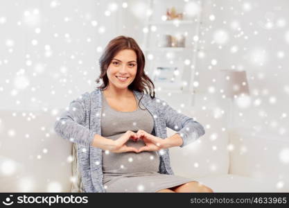 pregnancy, love, people, winter and expectation concept - happy pregnant woman sitting on sofa and making heart gesture at home over snow