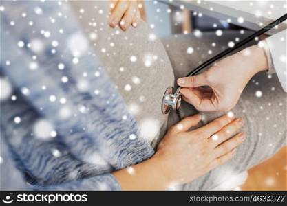 pregnancy, medicine, healthcare, winter and people concept - close up of gynecologist doctor with stethoscope listening to pregnant woman baby heartbeat at hospital over snow