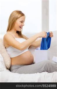 pregnancy, motherhood and celebration concept - smiling pregnant woman sitting on sofa and opening gift box with blue cardigan