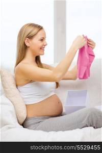 pregnancy, motherhood and celebration concept - smiling pregnant woman sitting on sofa and opening gift box with pink cardigan